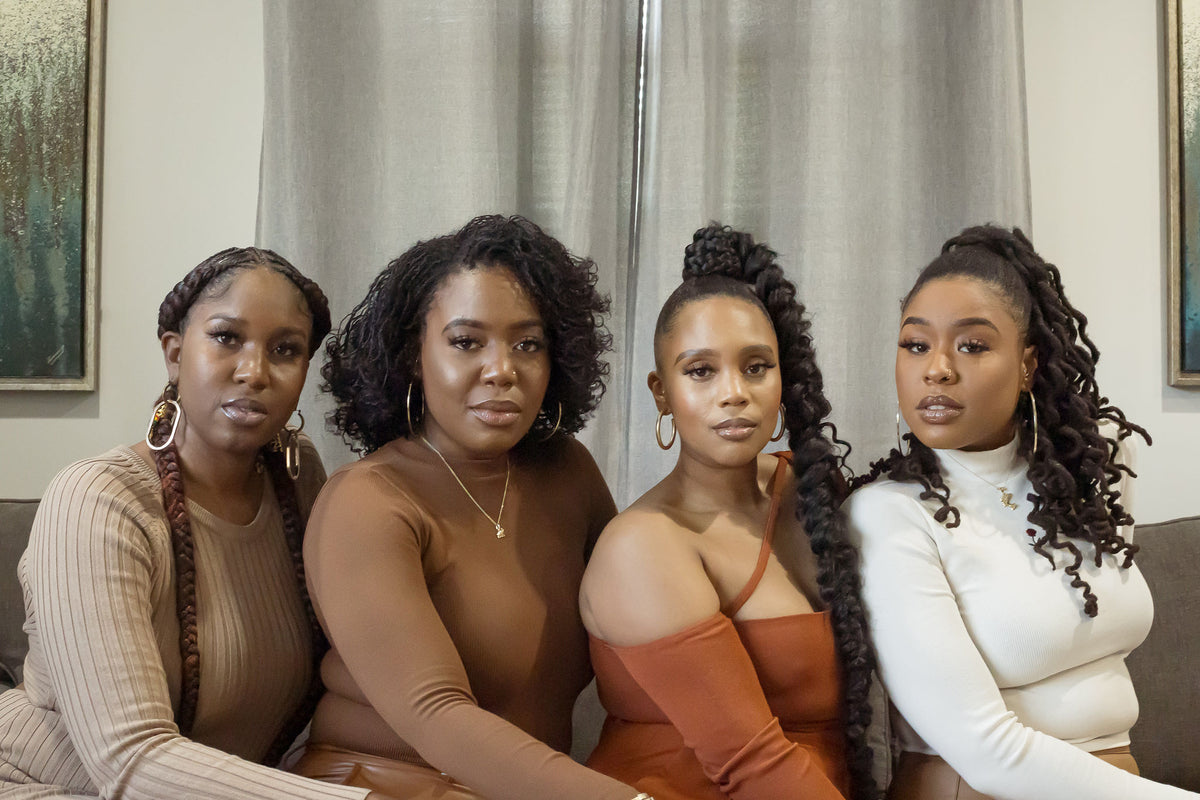 Four women sitting together indoors, wearing coordinated neutral-toned outfits including beige, brown, rust orange, and white. They are posed in front of light gray curtains with framed wall art visible in the background.